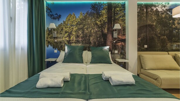A hotel room with a bed, two nightstands, a lamp, a sofa, and a large window with a forest and lake view. - Hotel Ciudad del Mar (Las Palmas de Gran Canaria)
