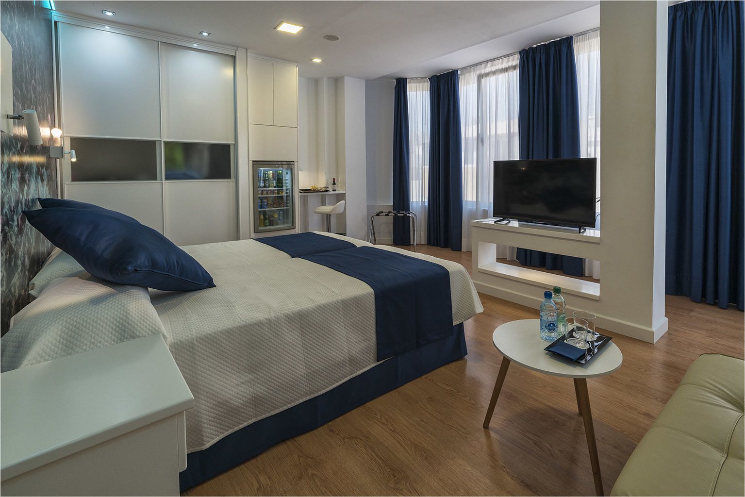 Una habitación de hotel moderna con cama, televisión, una mesita con una botella y vasos, y un minibar.