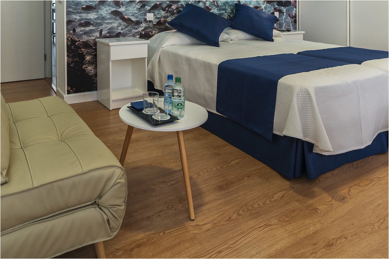 Quarto de hotel com uma cama, uma mesinha com um copo e uma cadeira.