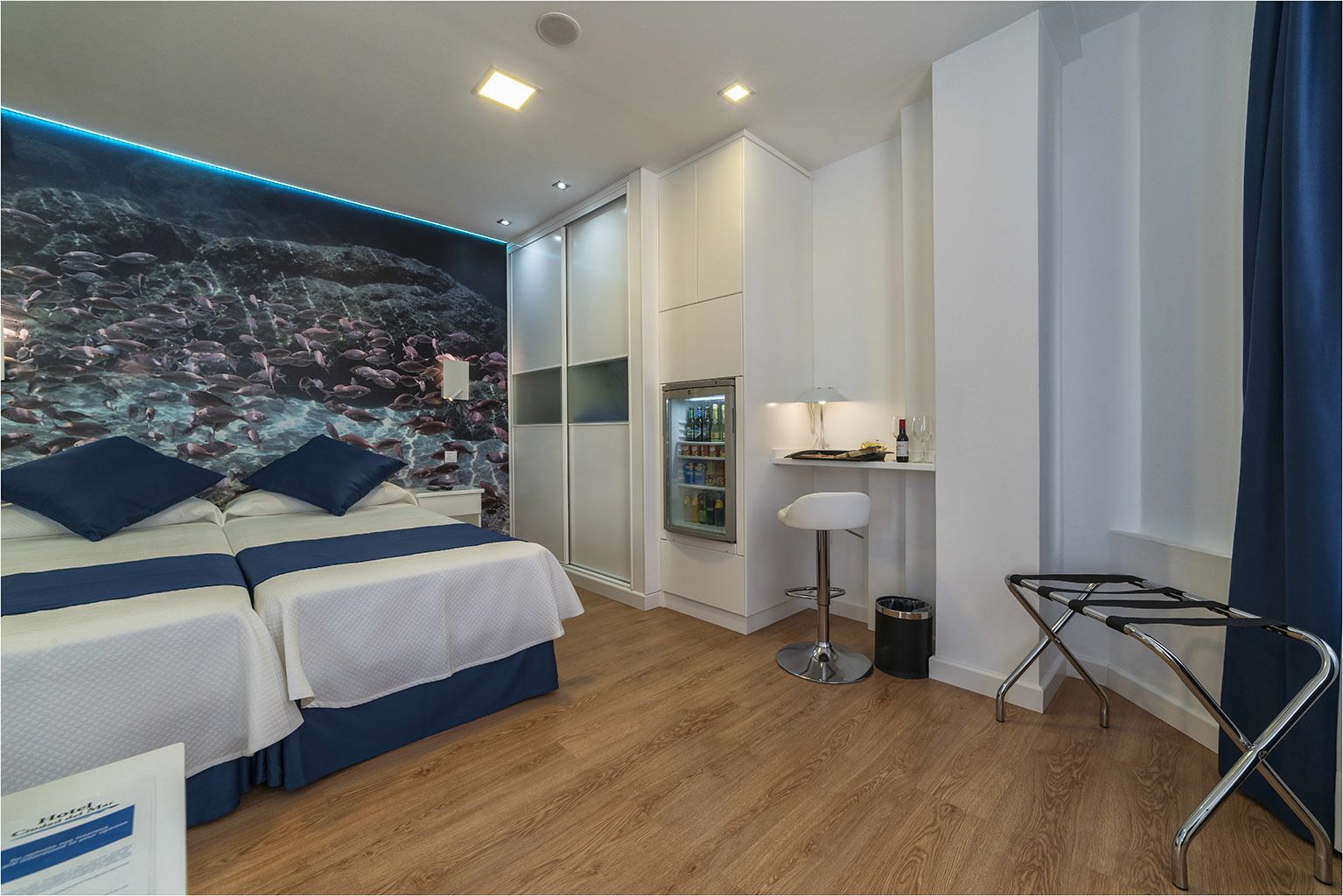 Une chambre d’hôtel avec un lit, un minibar, une petite table avec une lampe et une fenêtre avec des rideaux.