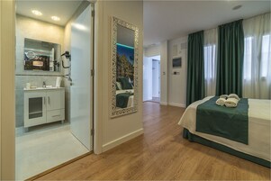 A hotel room with a wooden floor, a bed with a green bedspread, a bathroom with a sink and mirror, and a hallway with a door leading to another room.