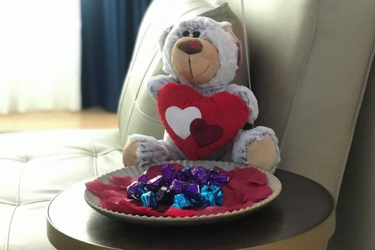 A plush toy bear holding a heart, placed on a cushion with a plate of wrapped gifts underneath.