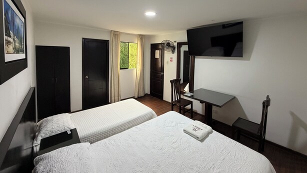 A hotel room with two beds, a desk, a chair, a TV, and a window with curtains. - Hostal Anthony's (Lima)