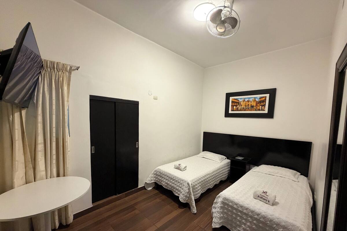 A hotel room with two beds, a small table, a ceiling fan, and a framed picture on the wall.