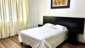 Cama bem arrumada com colcha branca, cabeceira escura, mesa de cabeceira, quadro na parede e janela com cortinas.