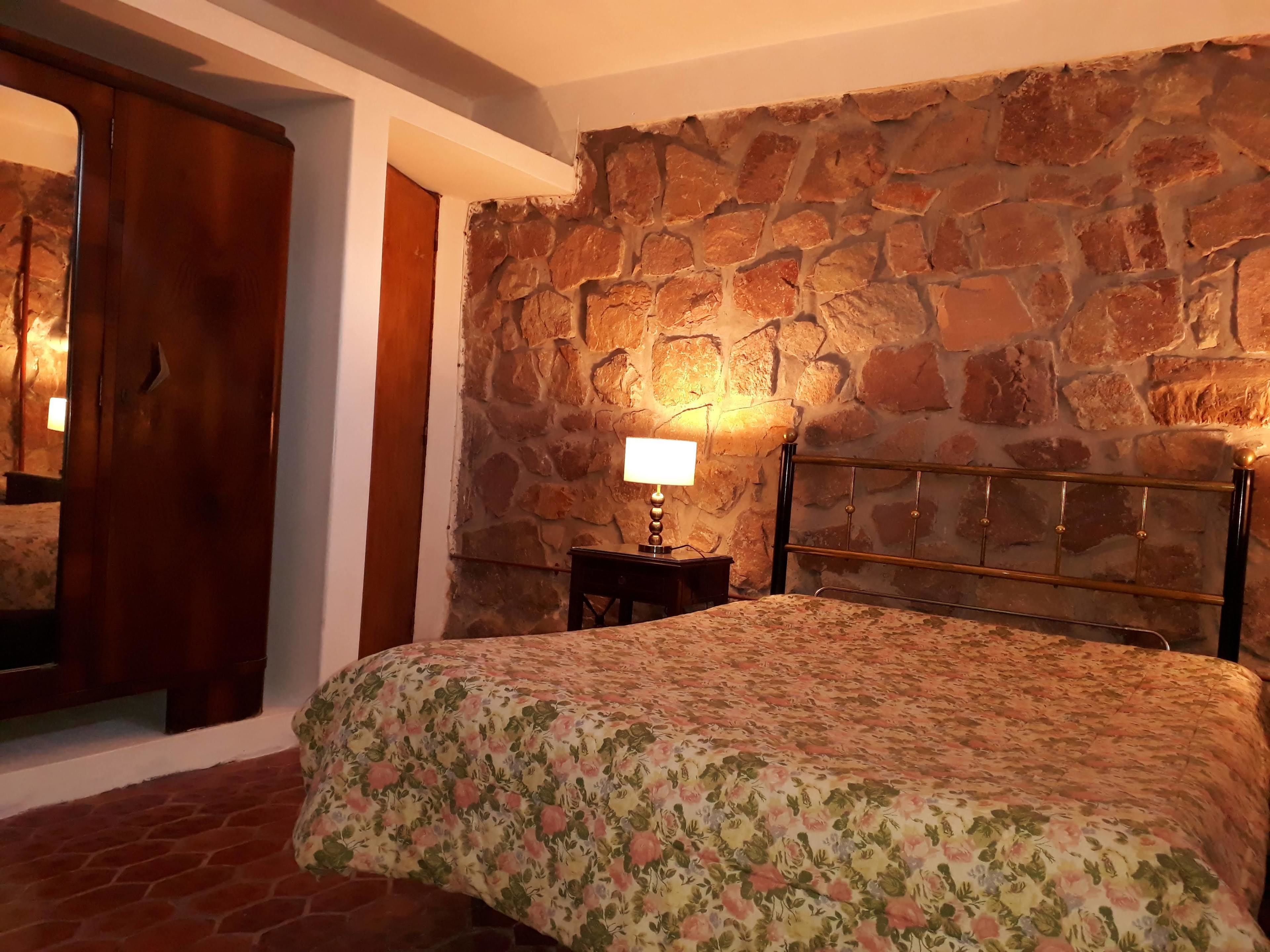 A bedroom with a stone wall, a wooden wardrobe, a bedside table with a lamp, and a floral bedspread.