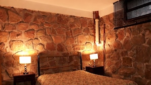 Une chambre avec un mur en pierre, un lit avec une literie à motifs floraux, deux tables de chevet avec des lampes et une tête de lit en bois.