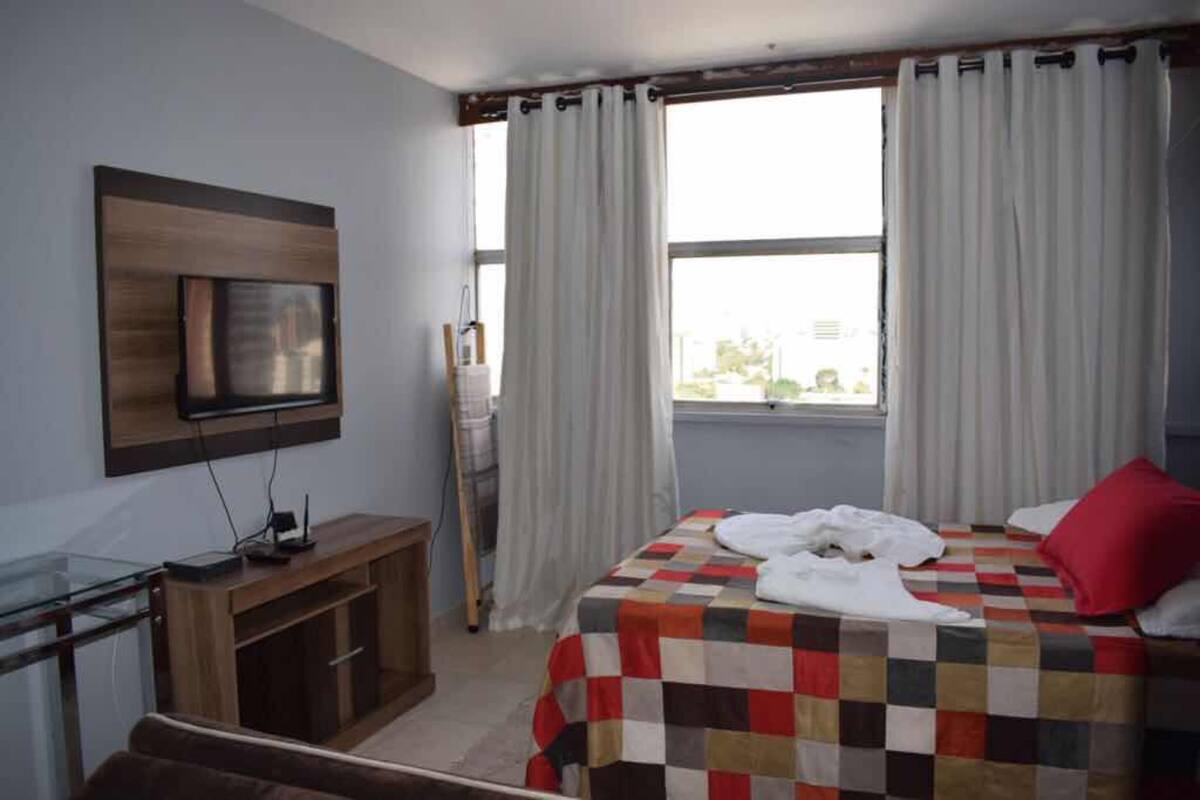 A hotel room with a bed, a TV mounted on the wall, a desk with a computer, and a window with curtains.