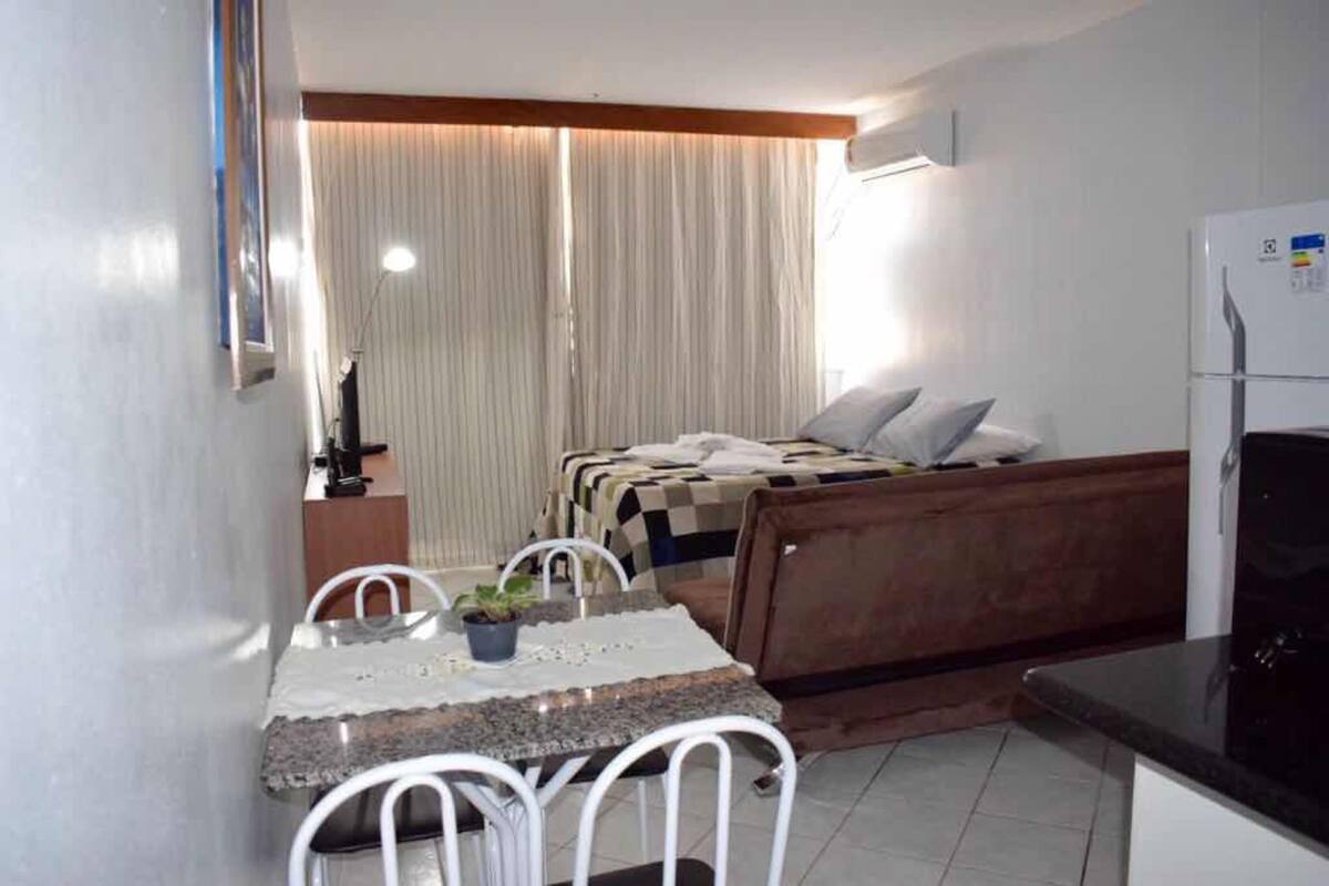 A compact hotel room with a kitchenette, a bed with a checkered blanket, a small dining table with chairs, and a potted plant.