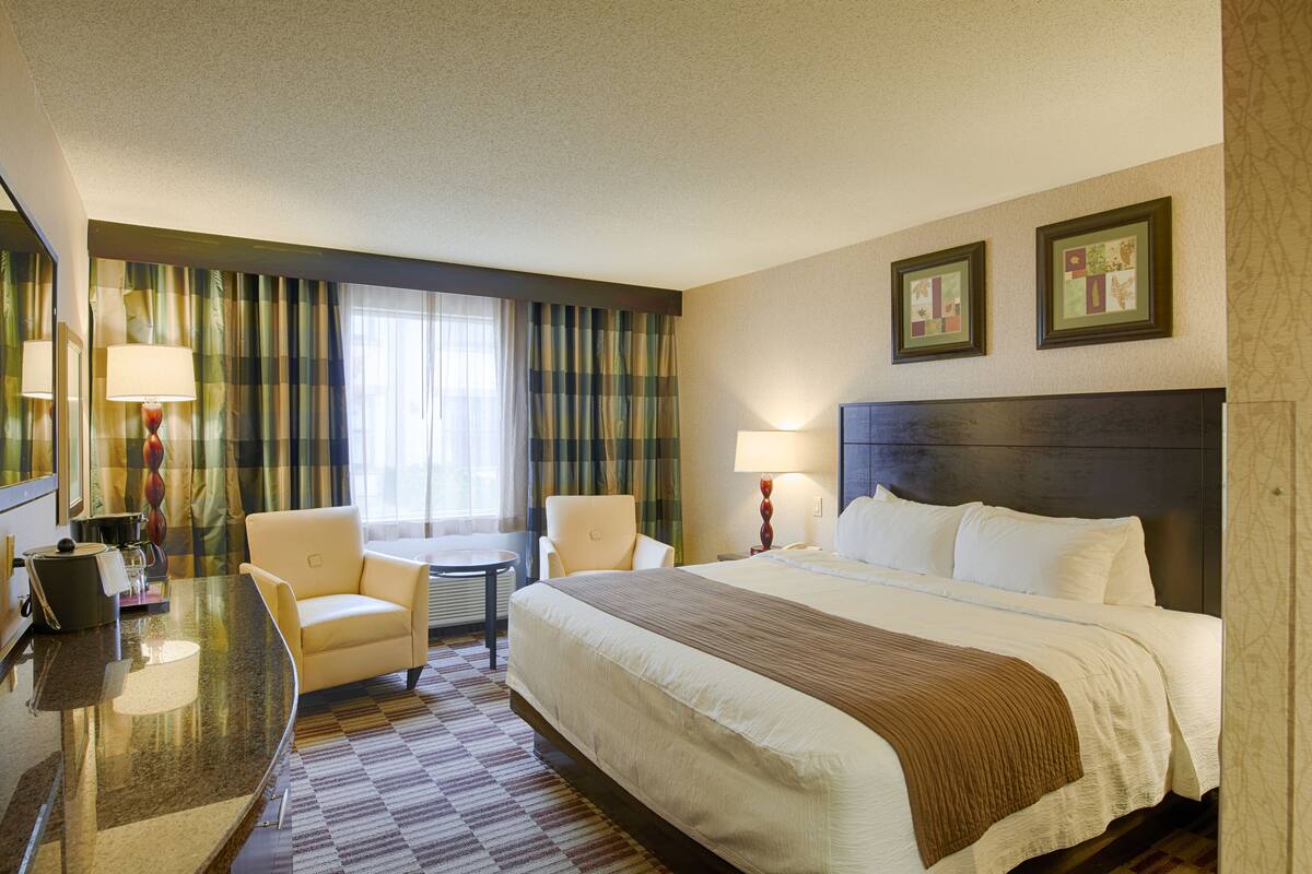 Ein Hotelzimmer mit einem Bett, zwei Sesseln, einem Schreibtisch und einem fenster mit Vorhängen.