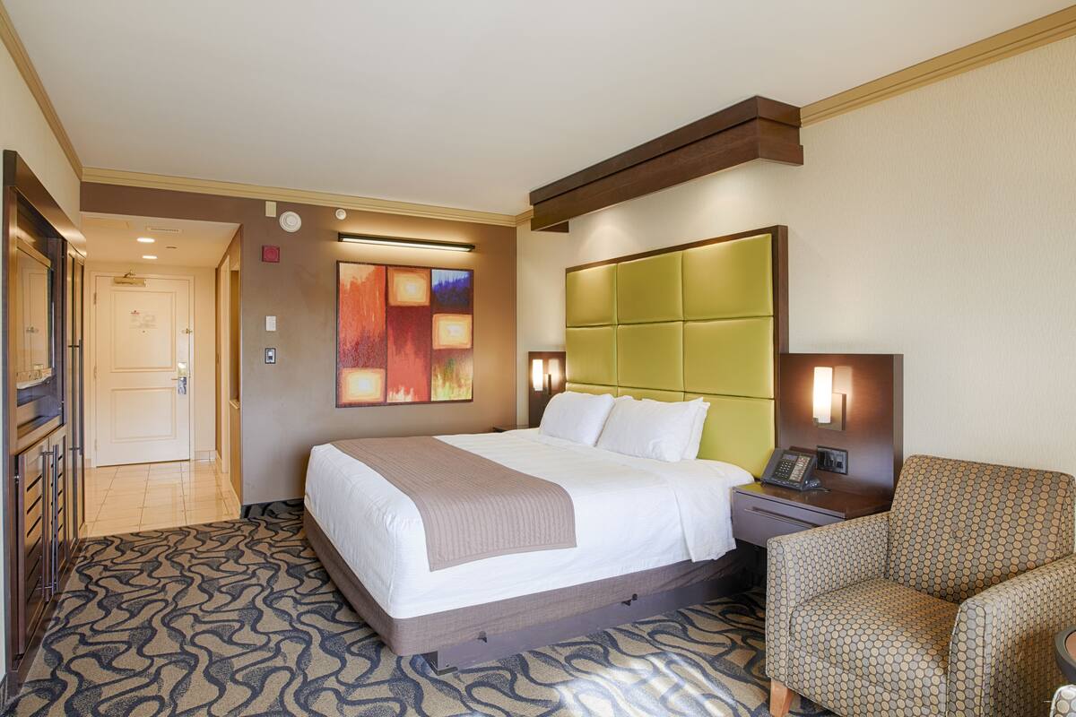Ein Hotelzimmer mit einem großen Bett, einem grünen Kopfteil, einem gemusterten Sessel und einem an der Wand montierten Telefon.
