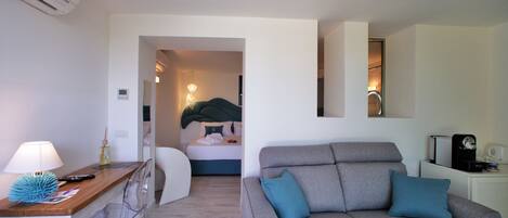 A modern living room with a grey sofa, a wooden table with a lamp, and a bed in the background.