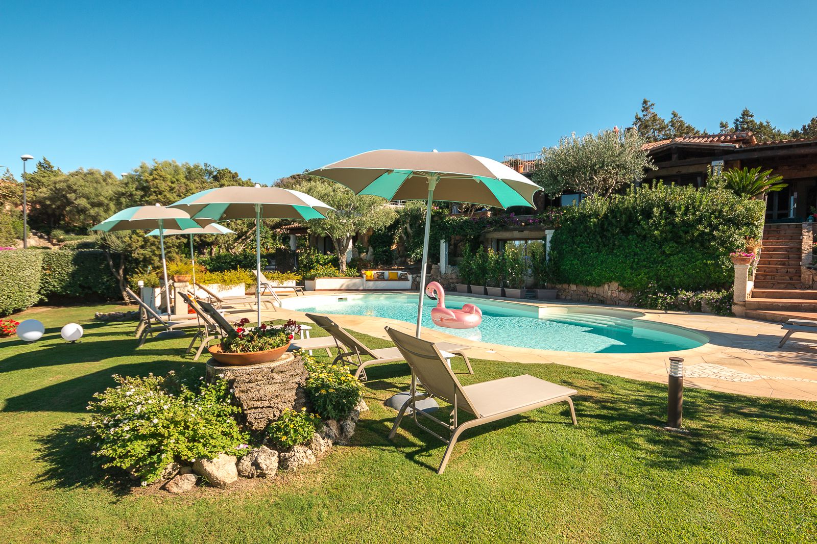 Piscina con sillones de descanso y sombrillas, rodeada de vegetación y una estructura de piedra.