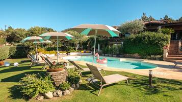 Piscina con sillones de descanso y sombrillas, rodeada de vegetación y una estructura de piedra.