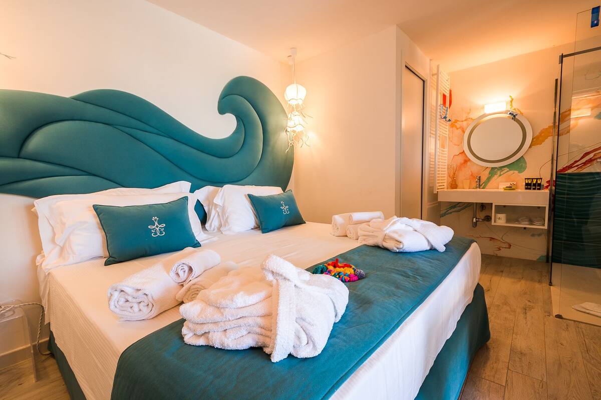 Een hotelkamer met een groot bed, een blauwgroen hoofdeinde, wit beddengoed, en een badkamer met een spiegel en handdoekrek.