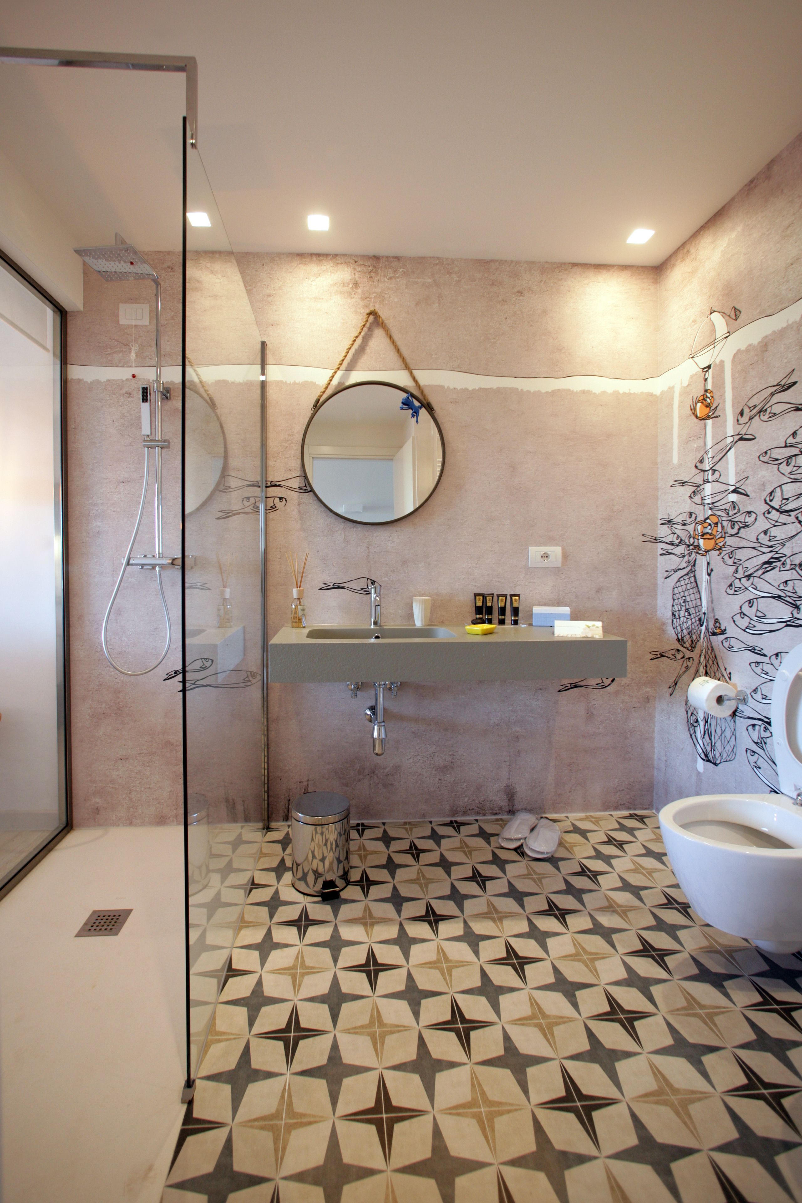 Een moderne badkamer met een vloer met een geometrisch patroon, een glazen douchecabine, een wastafel met spiegel erboven en een toilet met een decoratieve douchegordijn.