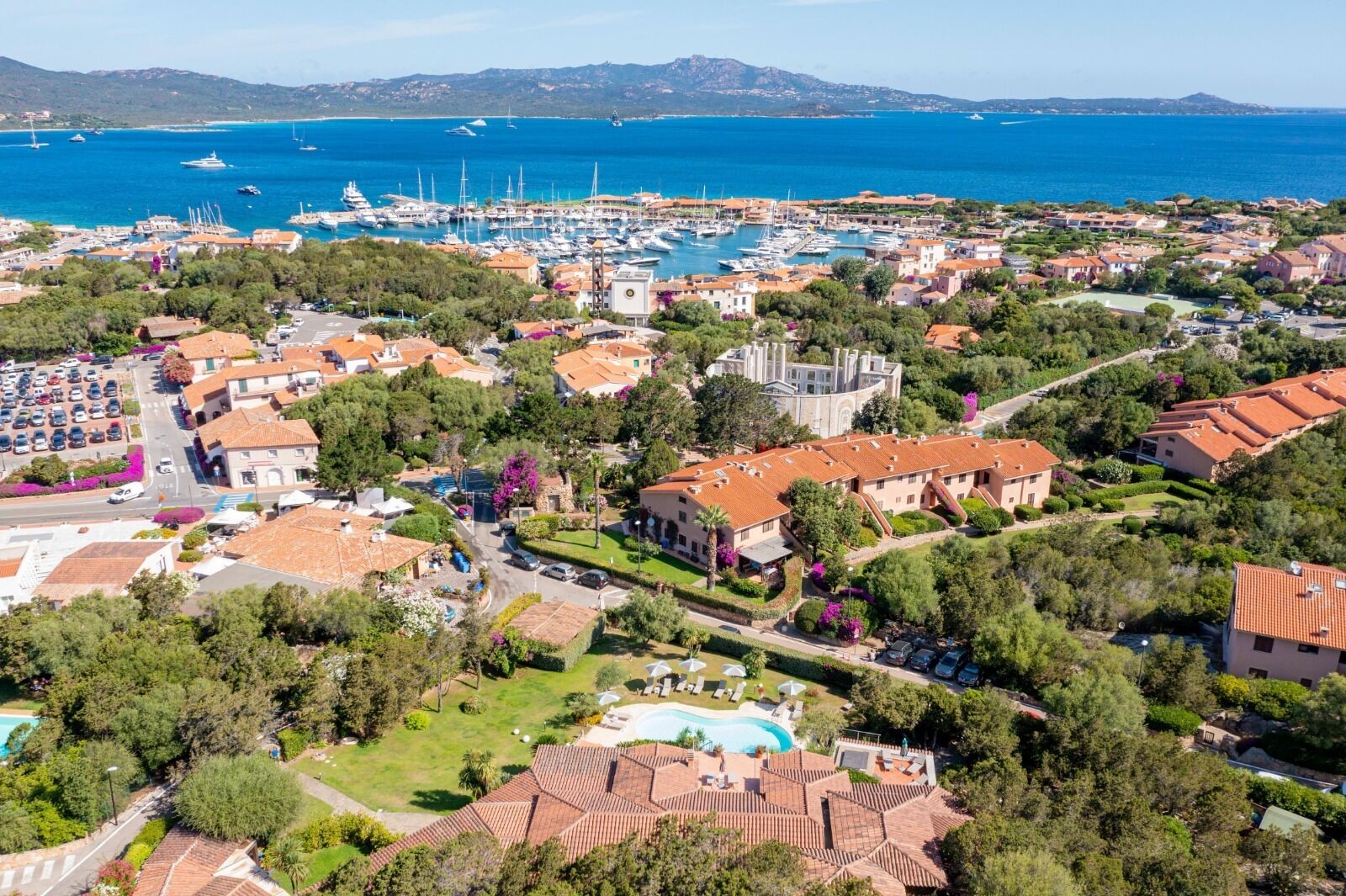 Vista aérea de un pueblo costero con un puerto deportivo, edificios residenciales y una piscina.