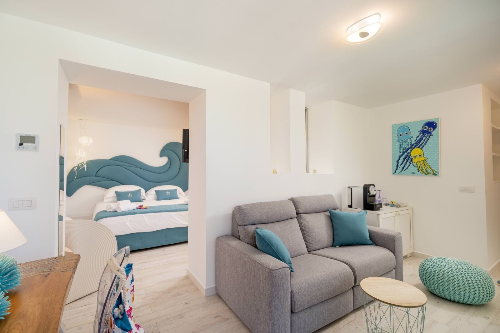 Ett modernt hotellrum med en grå soffa, en säng med ett vågmönstrat sänggavel, ett litet runt bord och en tavla med maneter på väggen.