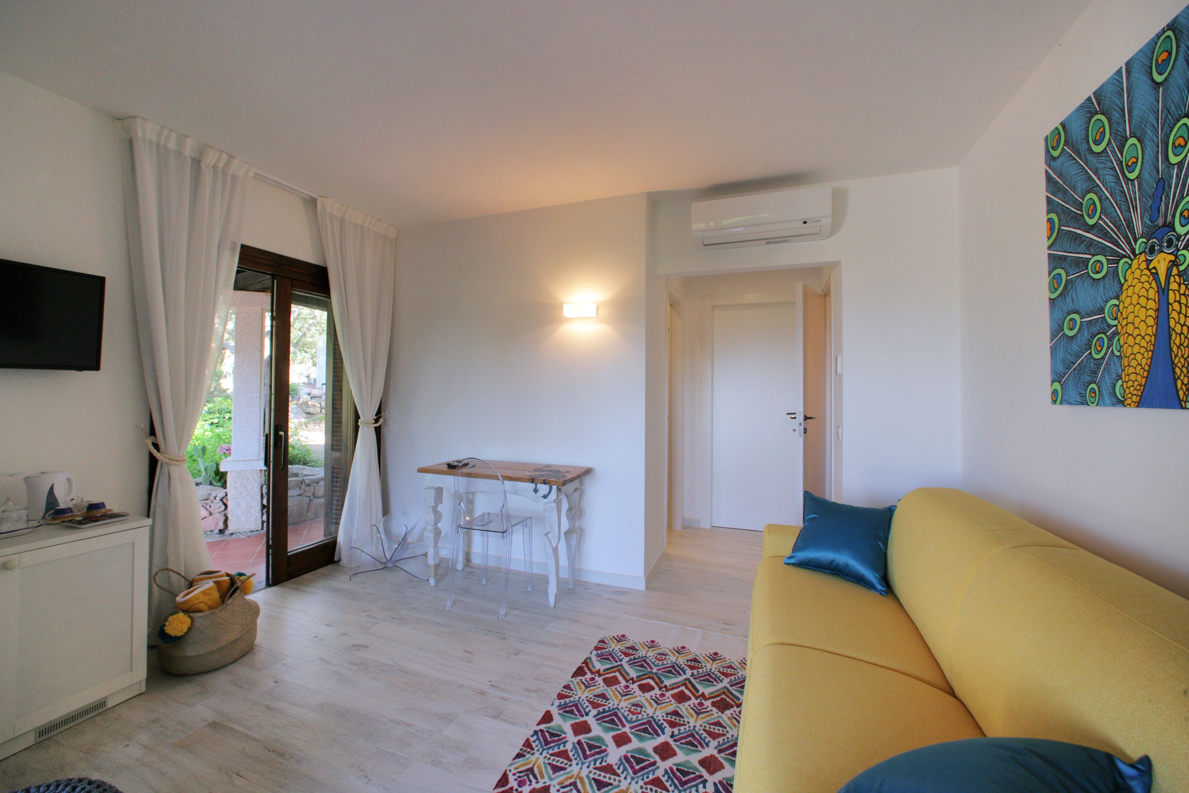 Un salon avec un canapé jaune, un tapis à motifs, une petite table et un tableau représentant un paon accroché au mur.