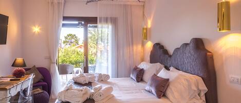 A hotel room with a bed, a desk with a lamp, and a view of greenery outside.