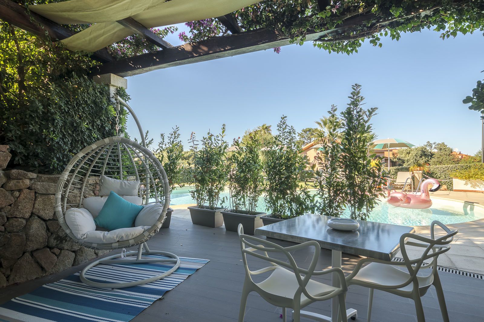 Eine Terrasse mit Korbstuhl, Tisch und Stühlen mit Blick auf einen Pool, in dem ein Schwan-Schwimmtiermodell schwebt.