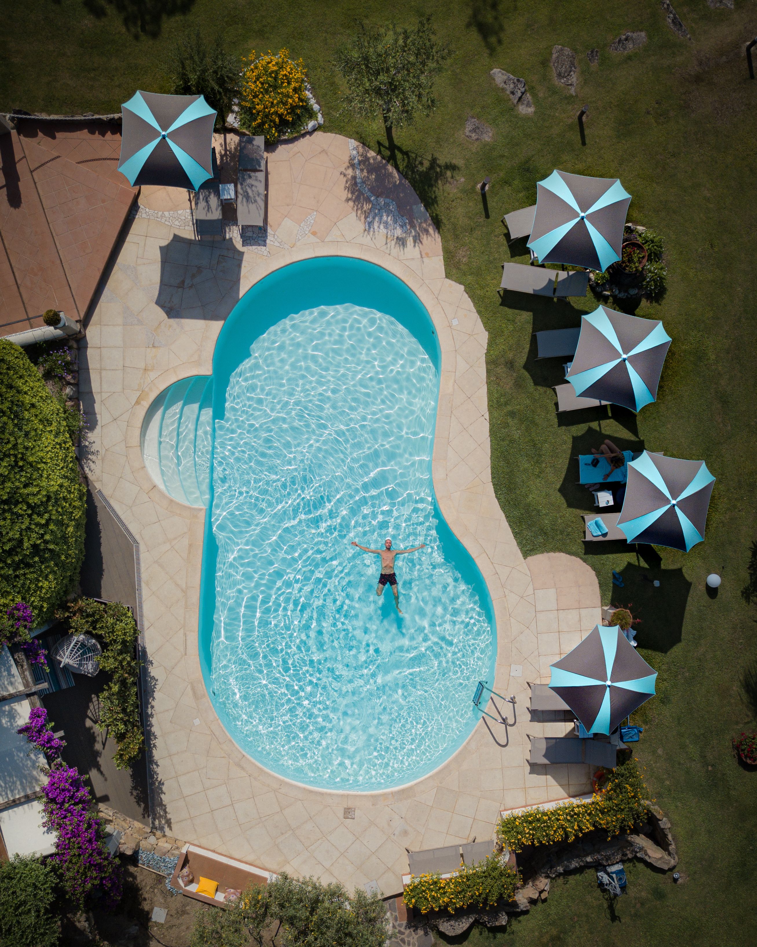 An aerial view of a swimming pool with a person in the water, surrounded by lounge chairs and umbrellas.