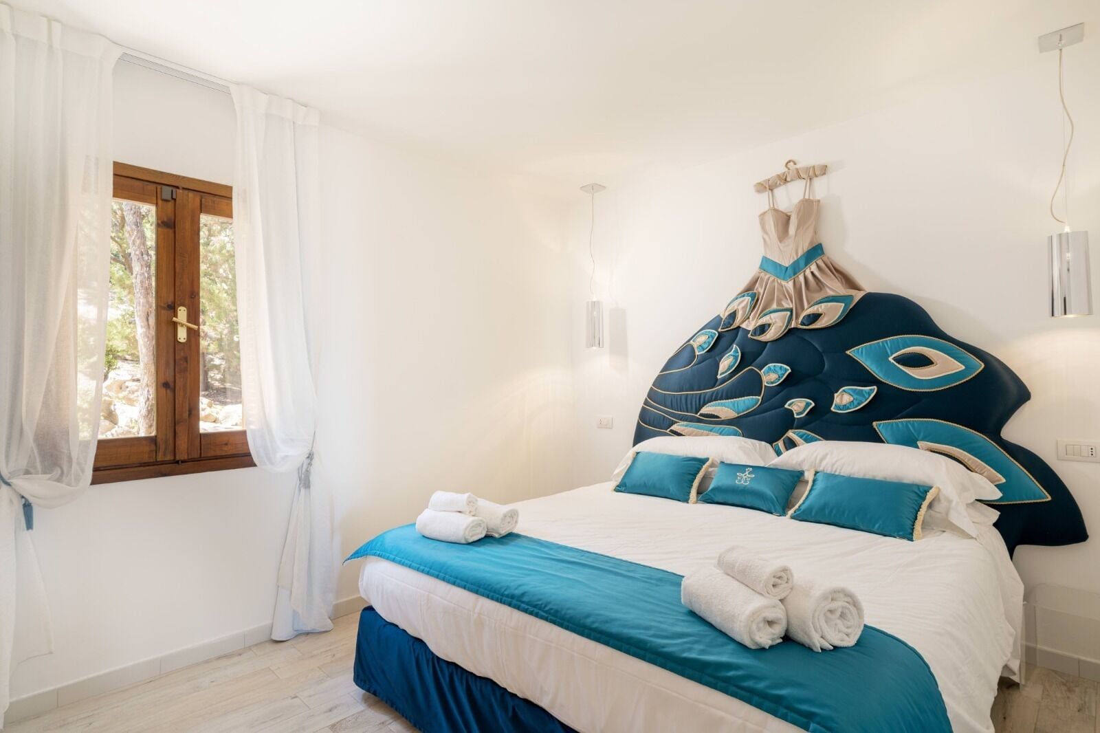 Un dormitorio con cama, cortinas blancas, una ventana y cabecera con almohadones decorativos.