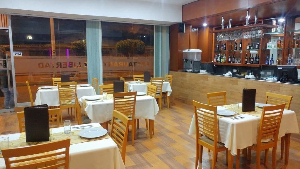 Un restaurante con varias mesas y sillas, una zona de bar con estantes llenos de botellas, y vistas amplias a través de grandes ventanales.