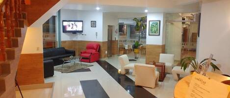 A hotel lobby with a staircase, a flat-screen TV, a reception desk, and a dining area.