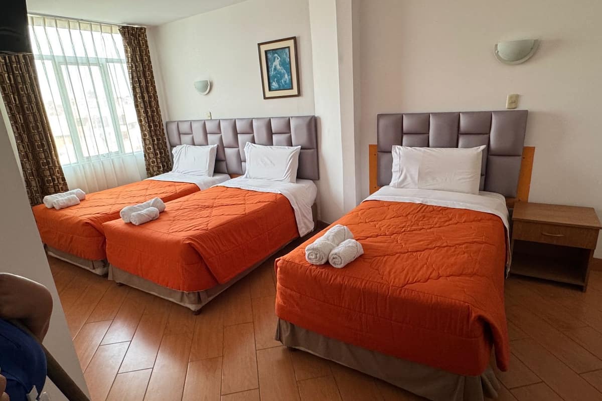 A hotel room with two beds, each with an orange bedspread and white pillows. There is a painting on the wall, a window with curtains, and a bedside table with a lamp.