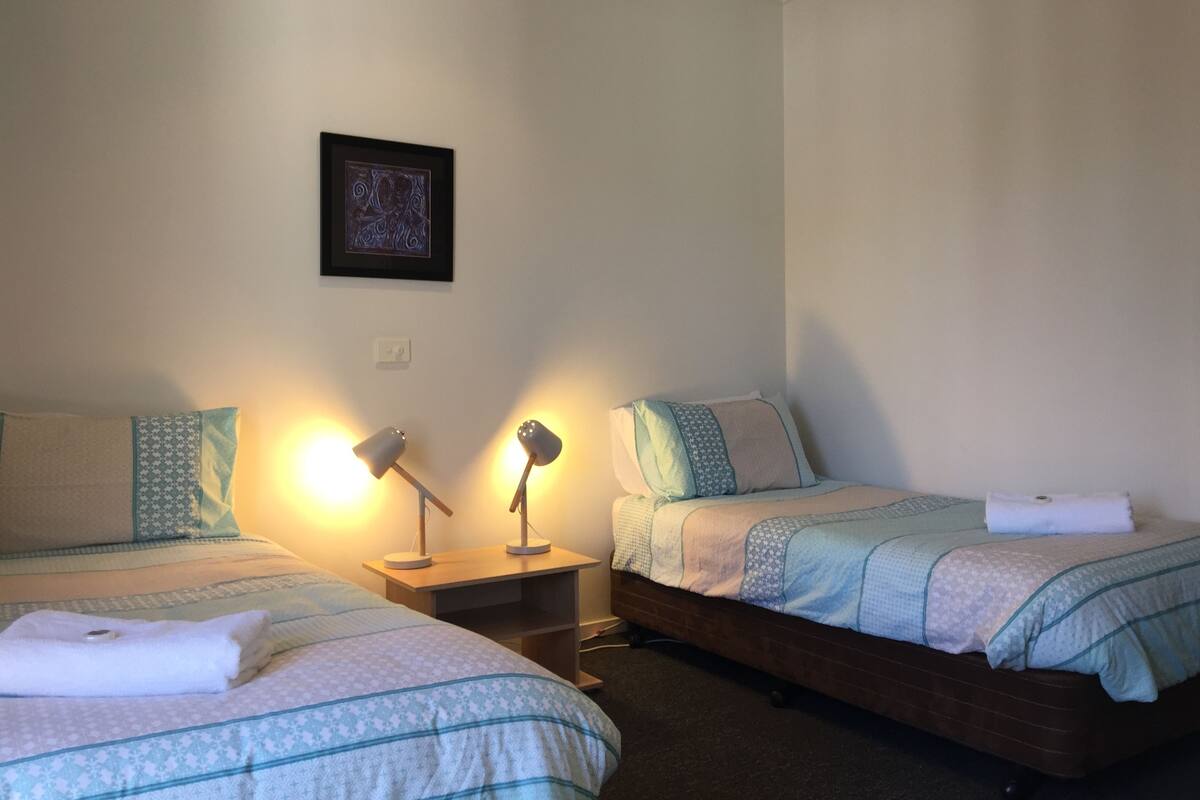 A bedroom with two beds, each with a patterned bedspread and pillows, a bedside table with lamps, and a framed picture on the wall.