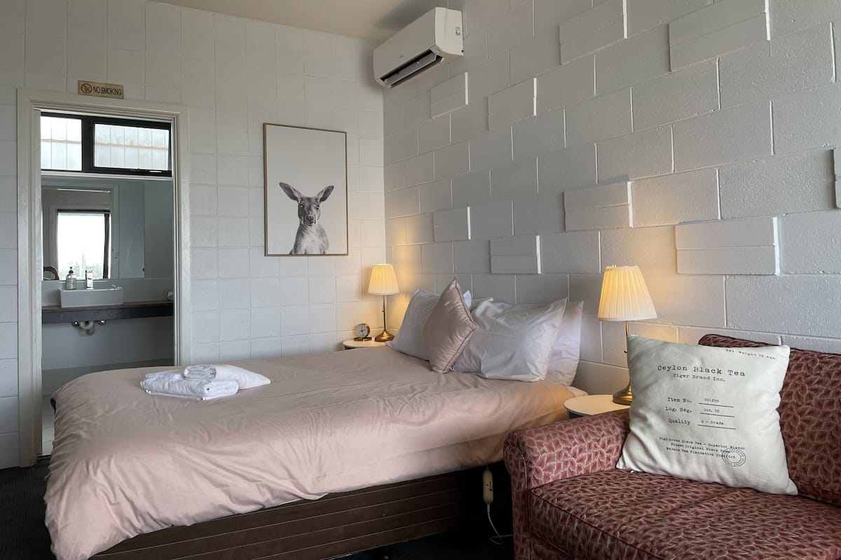 A hotel room with a bed, a bedside table with a lamp, a wall-mounted air conditioner, a no-smoking sign, a framed picture on the wall, and a window.