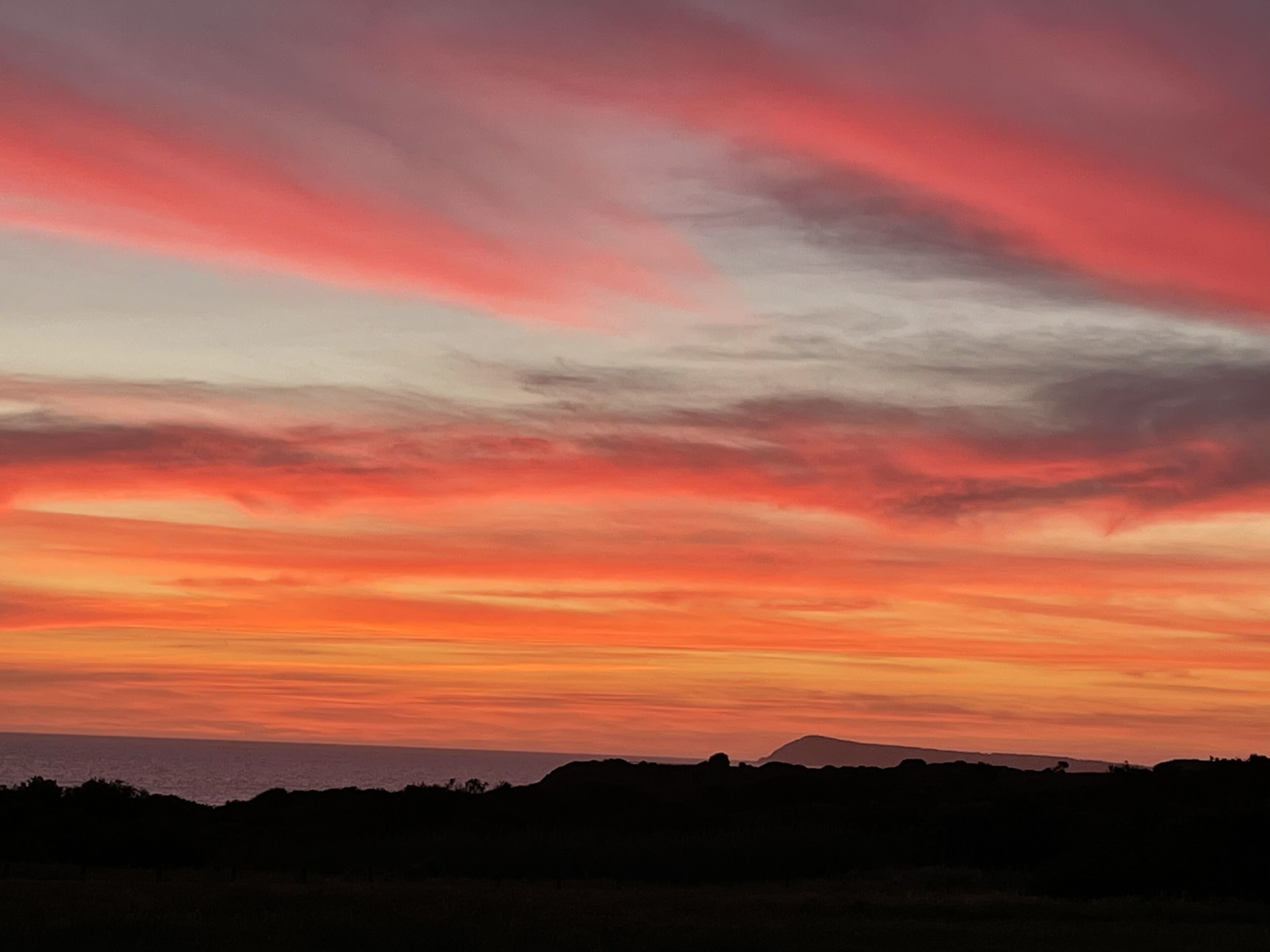 A sunset over a coastal landscape with silhouettes of hills and trees.