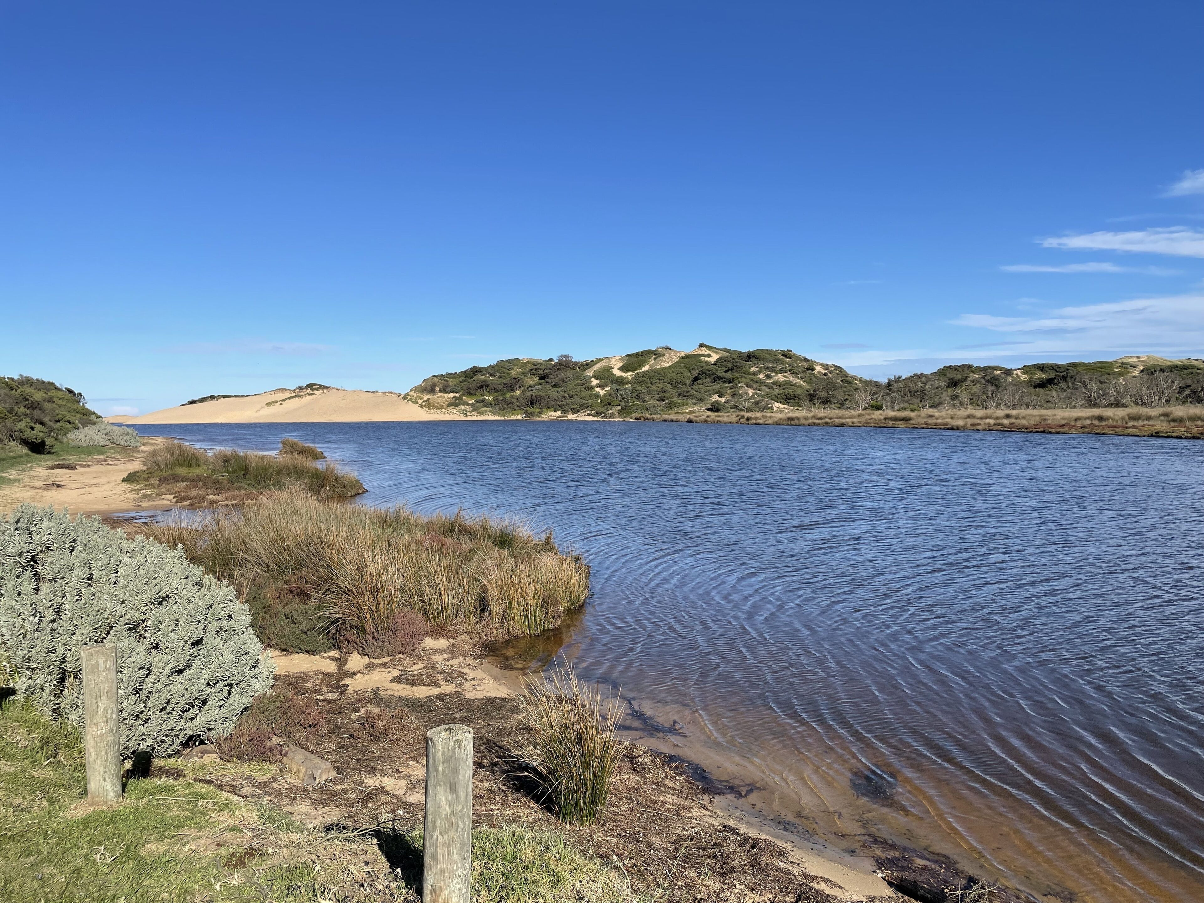 A river with sandy banks and grassy areas, surrounded by hills with sparse vegetation.