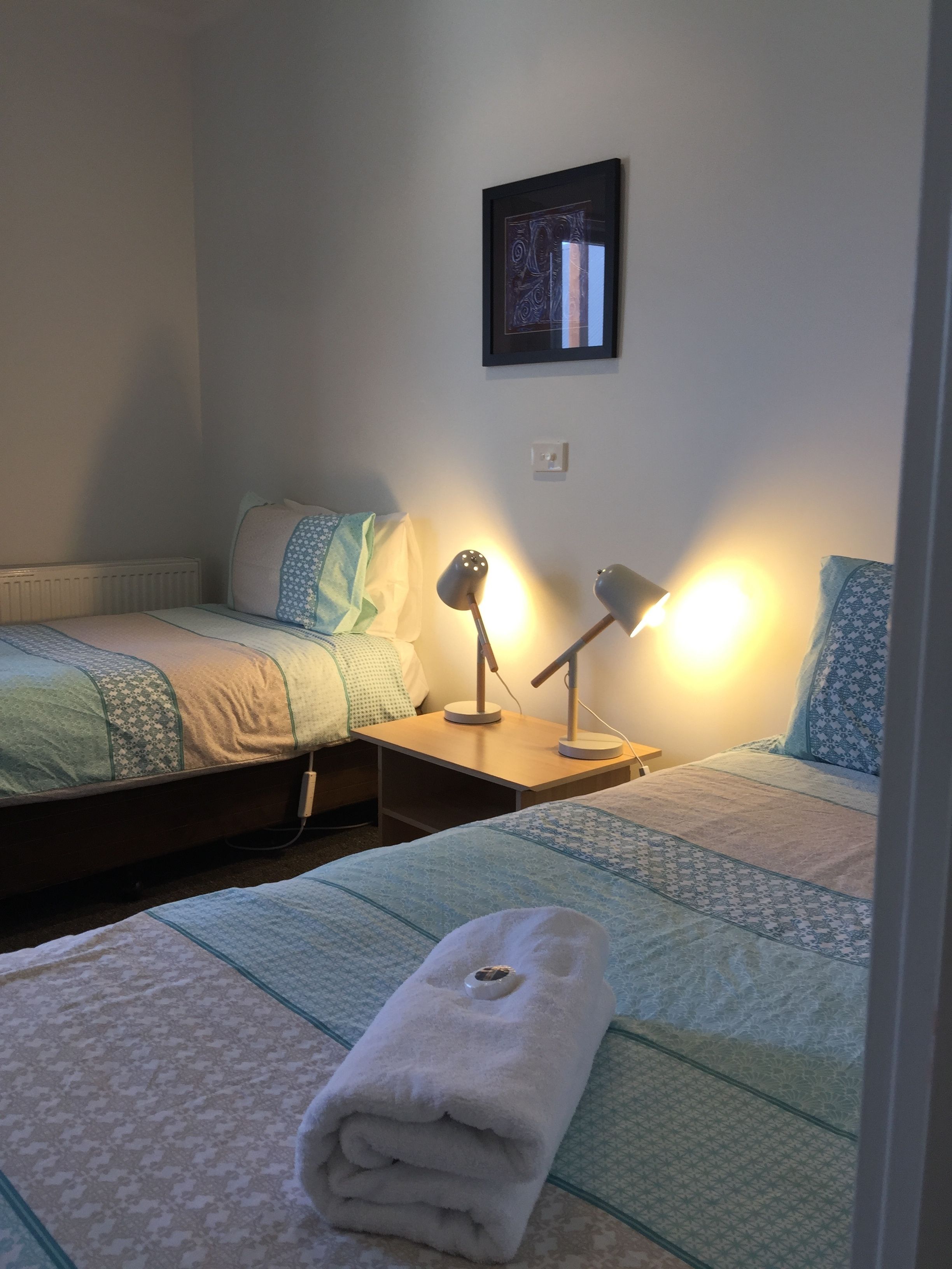 Een slaapkamer met twee bedden, elk met een bedspread en kussens met een patroon. Er staan twee nachtkastjes met lampen, en aan de muur hangt een ingelijst schilderij.