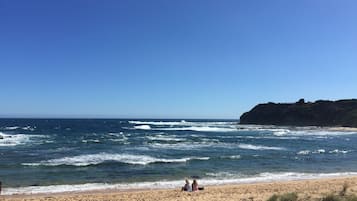 A beach with two people sitting on the sand, waves crashing onto the shore, and a rocky cliff in the background.