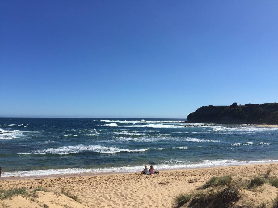 Une plage où deux personnes sont assises sur le sable, les vagues déferlant sur le rivage, et une falaise rocheuse en arrière-plan.