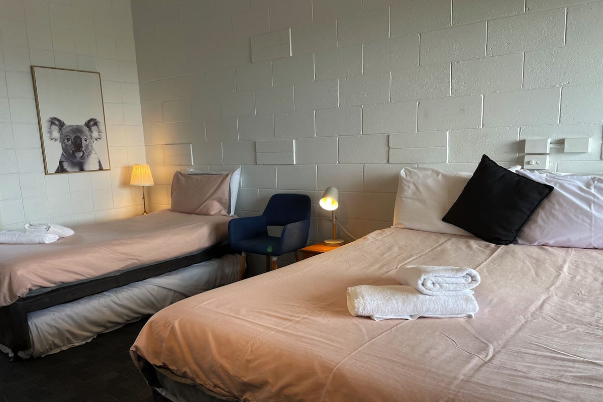 A hotel room with two single beds, a chair, a desk, a lamp, and a framed picture on the wall.
