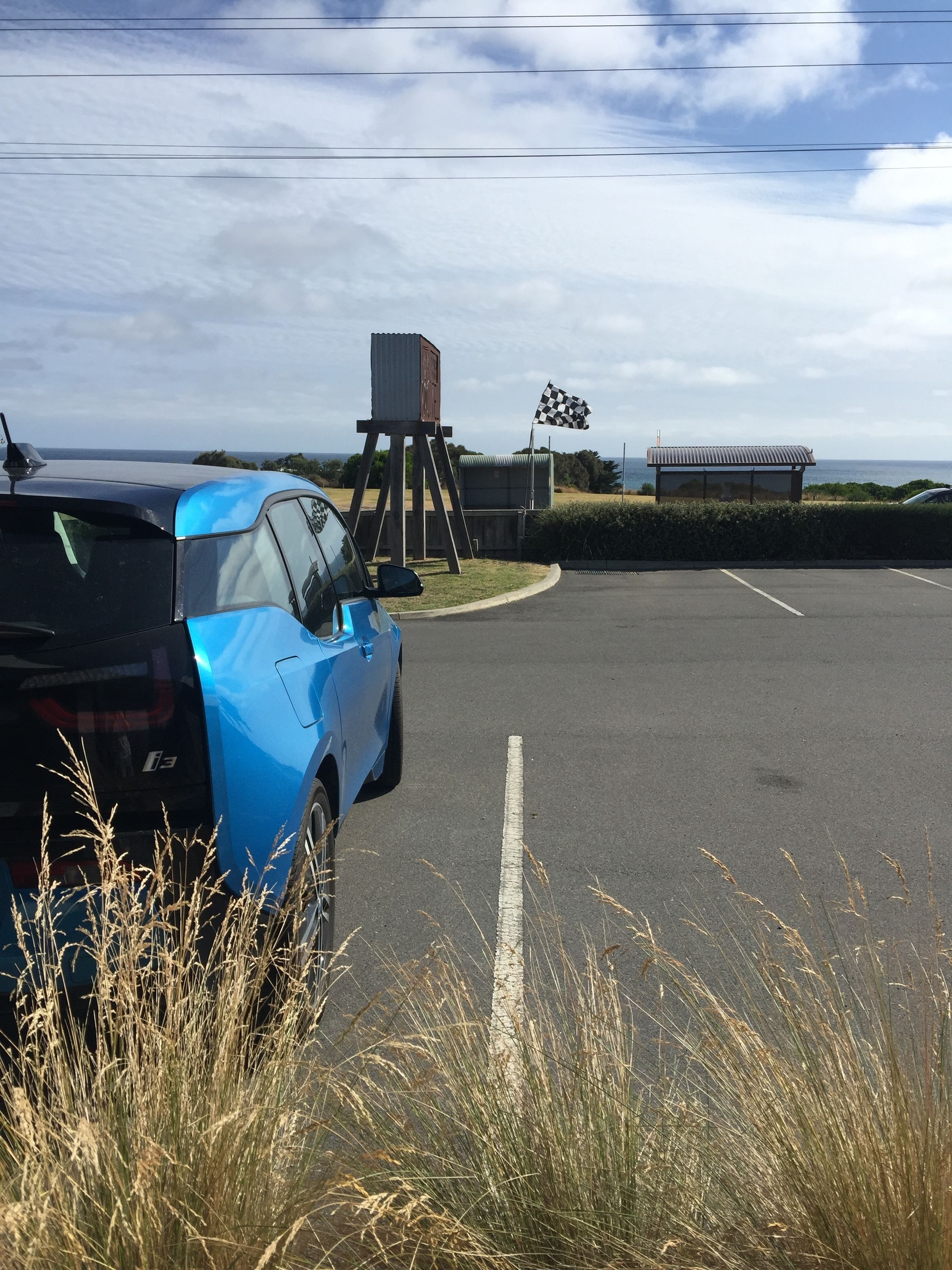A parking area with a blue car and a black car, a checkered flag, and a small building with a metal roof.