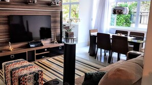 Uma sala de estar moderna com televisão de tela plana, um espaço de refeições com mesa e cadeiras, e uma grande janela com vista para o verde.