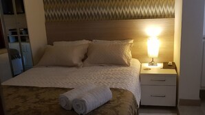 Cama bem arrumada com cobertura acolchoada, dois travesseiros e uma toalha enrolada ao lado. Há uma mesa de cabeceira com uma luminária e um pequeno objeto decorativo. O quarto possui papel de parede estampado e cabeceira de madeira.