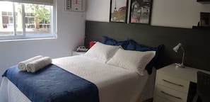 Un lit bien fait, avec une literie blanche et bleue, une table de chevet avec une lampe et une fenêtre donnant sur l’extérieur.