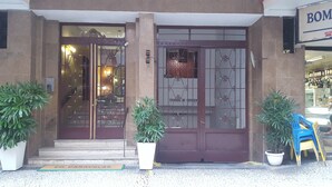 Entrada de hotel com porta marrom escura, placa de entrada de serviço e plantas em vasos.