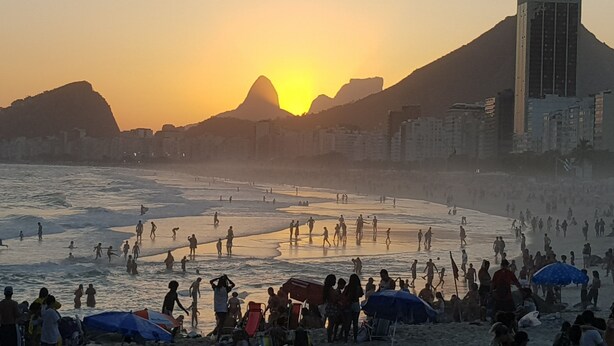 Cena de praia com pessoas e guarda-sóis, uma montanha ao fundo e edifícios ao longo da costa.