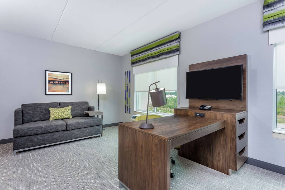 A modern office space with a grey sofa, a wooden desk, a flat-screen TV, and a window with a view.