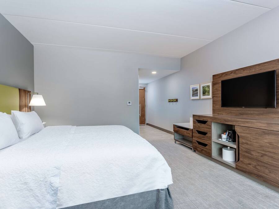 Una habitación de hotel moderna con una cama grande, un televisor de pantalla plana montado en un mueble de madera y un pasillo que conduce a otra habitación.