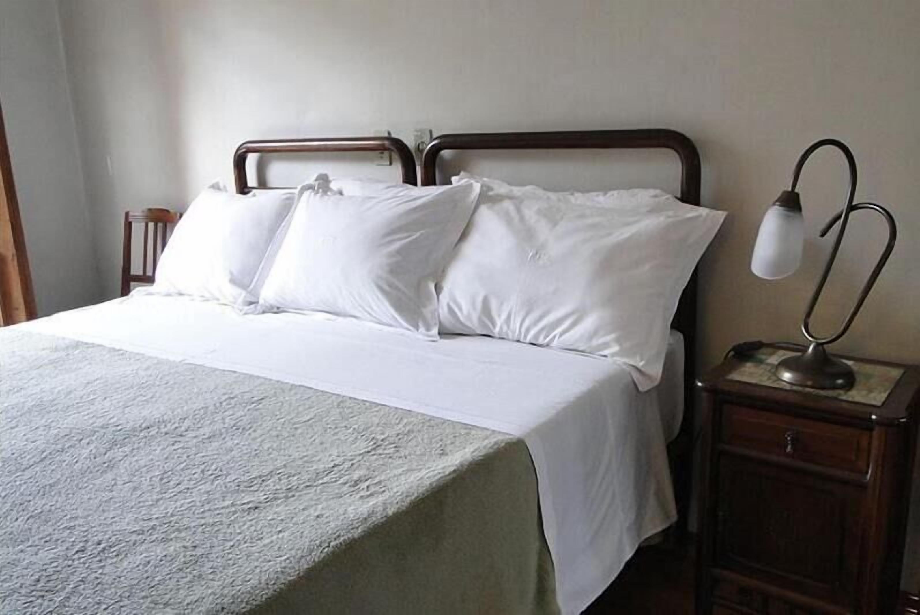 Uma cama bem arrumada com lençóis brancos, cabeceira de madeira e mesa de cabeceira com abajur.
