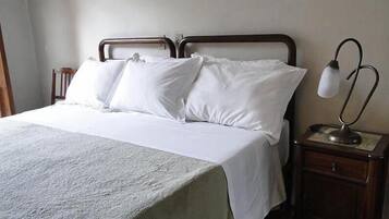 Uma cama bem arrumada com lençóis brancos, cabeceira de madeira e mesa de cabeceira com abajur.