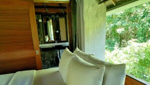 Una cama con sábanas y almohadas blancas, una pared de piedra, una puerta de madera y una ventana con vistas al verdor.