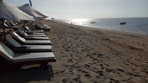 Una playa con tumbonas y sombrillas, un bote en el agua, e huellas en la arena.